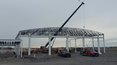 Crane lowering a bowstring joist onto beam structure at airport hangar construction site.